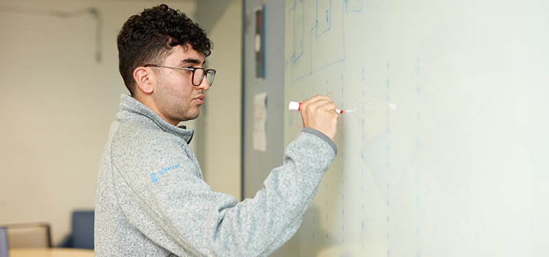 Young man with glasses standing at white board working out a problem